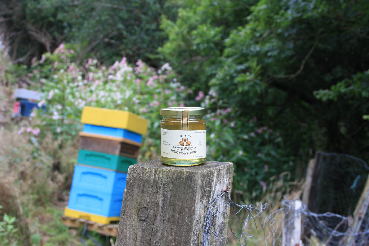 Jar of honey on a wooden post with beehives and flowers in the background