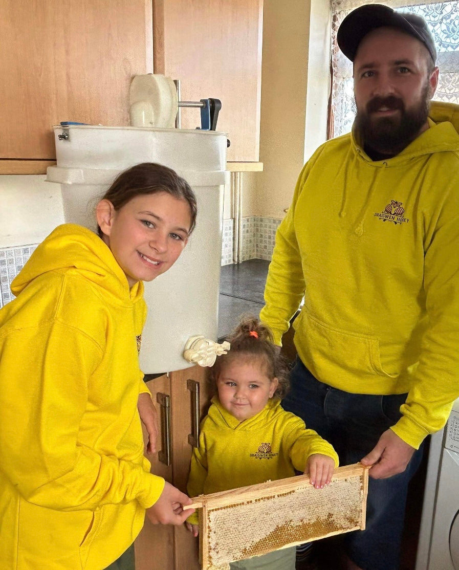 a father and two daughters wearing matching yellow hoodies holding a frame of honey during their first ever honey extraction